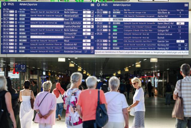 11 July 2026, North Rhine-Westphalia, Duesseldorf: Travelers stand in front of the train information board at the main station, which displays delays and a notice about vandalism. According to Deutsche Bahn, the embankment between Langenfeld and Leverkusen caught fire in two places on Friday morning. The fire department extinguished the fires, but the fire caused damage to several signal cables, preventing trains from running on that section of the line. Photo: Christoph Reichwein/dpa