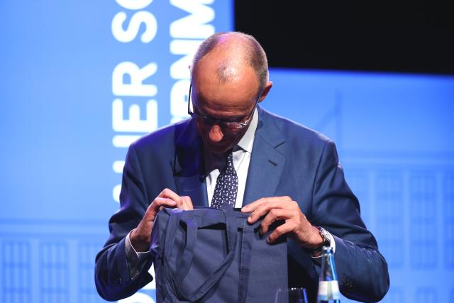 13 July 2026, North Rhine-Westphalia, Cologne: German Chancellor Friedrich Merz looks into a bag during the opening of the Adenauer School of Government (ASG) in Speyer, Germany. The new institution aims to train future decision-makers, with a discussion between the Chancellor and students scheduled to follow his speech. Photo: Rolf Vennenbernd/dpa