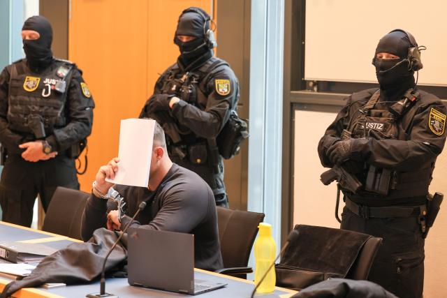 20 July 2026, Saxony-Anhalt, Magdeburg: The handcuffed defendant sits before court officers in a courtroom at the Regional Court. The resident of Aschersleben faces charges of attempted murder following a violent clash at a 2nd Bundesliga soccer match between 1. FC Magdeburg and SG Dynamo Dresden at Magdeburg Stadium. Prosecutors allege the man threw a 15-kilogram paving stone with full force at a police cordon. Photo: Peter Gercke/dpa