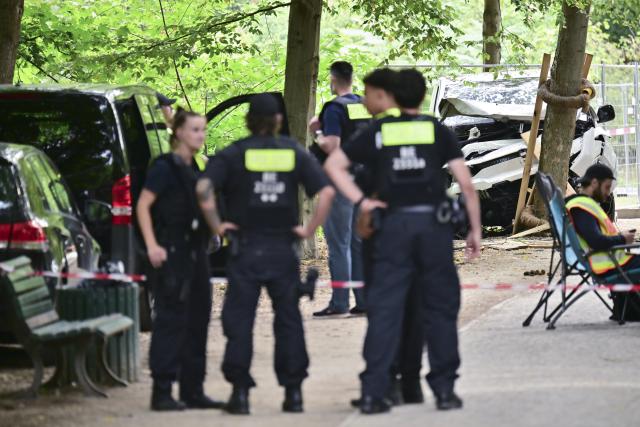 26 July 2026, Berlin: Police officers stand in front of a wrecked white vehicle in the Tiergarten area, where it had crashed into a tree. On the sidelines of Christopher Street Day in Berlin, the vehicle had plowed into a crowd. One woman was killed and 16 other people were injured, some seriously. Photo: Sebastian Gollnow/dpa