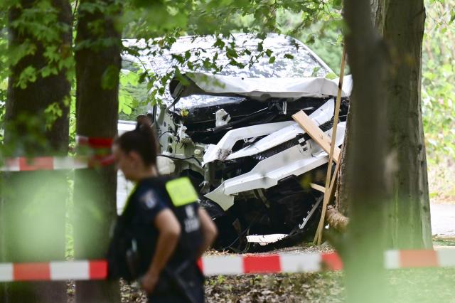 26 July 2026, Berlin: A police officer walks among the trees near the Tiergarten, passing a wrecked white vehicle that had crashed into a tree. On the sidelines of Christopher Street Day in Berlin, the vehicle had plowed into a crowd. One woman was killed and 16 other people were injured, some seriously. Photo: Sebastian Gollnow/dpa