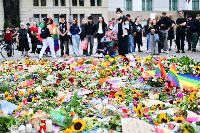 27 July 2026, Berlin: People standing around flowers, flags, and candles left in front of the Brandenburg Gate as a sign of condolence, following the terrorist attack on the sidelines of Christopher Street Day (CSD). Photo: Sebastian Gollnow/dpa
