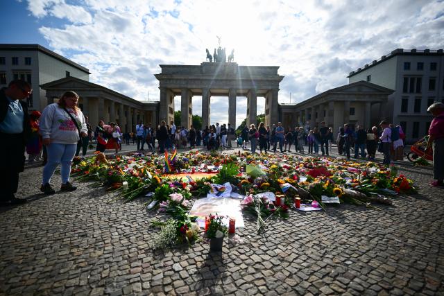 27 July 2026, Berlin: People standing around flowers, flags, and candles left in front of the Brandenburg Gate as a sign of condolence, following the terrorist attack on the sidelines of Christopher Street Day (CSD). Photo: Sebastian Gollnow/dpa