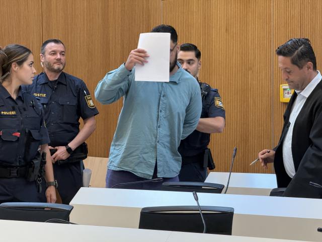28 July 2026, Bavaria, Munich: The defendant, Farhad N, covers his face as he stands in the courtroom next to his attorney Oemer Sahinci (R) and court officials. Farhad N. is charged with two counts of murder following a car attack on a demonstration in Munich on February 13, 2025. Photo: Elke Richter/dpa