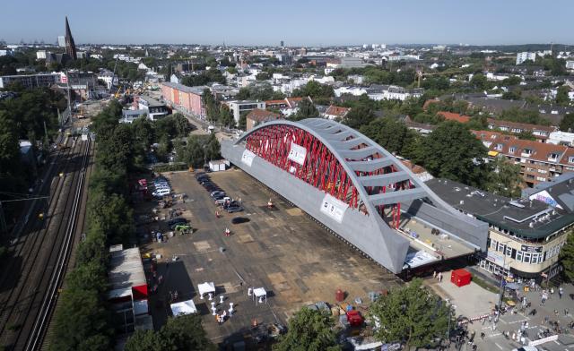 30 July 2026, Hamburg: A view shows the massive steel shell of the new Sternbruecke railway bridge positioned on the roadway of Max-Brauer-Allee. Crews successfully moved the 108-meter-long and 21-meter-wide, column-free arch-and-beam structure out of its assembly area. The complex transport of the bridge to its final destination at the intersection of Stresemannstraße and Max-Brauer-Allee is scheduled to continue through the day. Photo: Christian Charisius/dpa