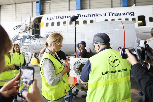31 July 2026, Hamburg: German Minister of Education Karin Prien speaks with Abu Bakr Chargui, an aircraft mechanic apprentice at Lufthansa Technik, in front of a decommissioned Boeing 737 in a maintenance hangar at Lufthansa Technik. Prien visited Lufthansa Technik on Friday and discussed opportunities in vocational training during a tour of the facility. Photo: Christian Charisius/dpa