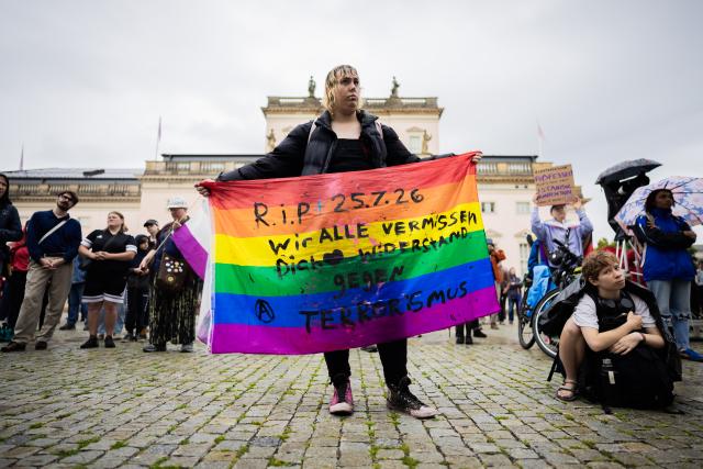 01 August 2026, Berlin: A person holding a rainbow flag with the inscription "R.I.P. - 7/25/26 - We all miss you - Resistance against terrorism" stands at a rally under the slogan "We will not be divided" in solidarity with the queer community following the attack on the outskirts of Christopher Street Day last week at Bebelplatz. Photo: Christoph Soeder/dpa