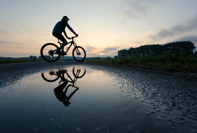 02 August 2026, Baden-Wuerttemberg, Riedlingen: A cyclist is reflected in a rain puddle as the sun rises in Baden-Wuerttemberg. Photo: Thomas Warnack/dpa