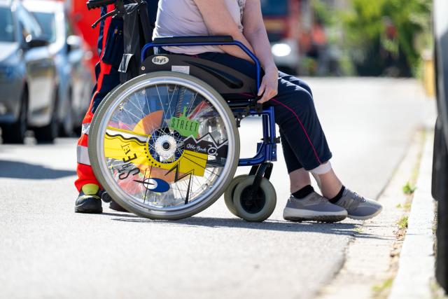 08 August 2026, Bavaria, Rednitzhembach: A woman in a wheelchair is being pushed across the street in front of a senior living complex in Roth County, after a fire has broken out at the  facility. The residents have been evacuated. Photo: Pia Bayer/dpa
