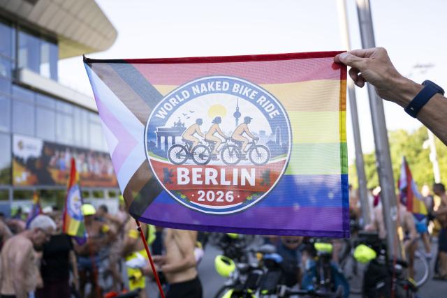 09 August 2026, Berlin: A participant in the "World Naked Bike Ride (WNBR) Berlin 2026" bike rally is holding a flag that reads "World Naked Bike Ride (WNBR) Berlin 2026." Through this event, the participants are advocating for bodily autonomy, mutual respect, and positive coexistence. Photo: Christophe Gateau/dpa