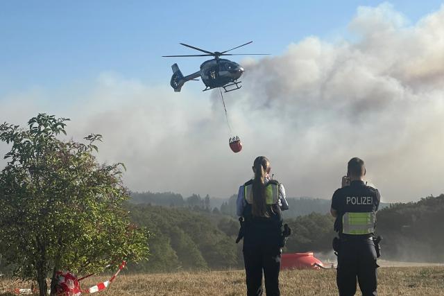 14 August 2026, North Rhine-Westphalia, Hürtgenwald: A police helicopter equipped with an external water tank takes off to assist in firefighting efforts at the forest fire in the Hürtgenwald. Several hundred hectares of forest are ablaze. Photo: Christoph Driessen/dpa