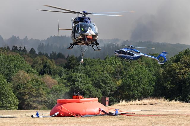 14 August 2026, North Rhine-Westphalia, Hürtgenwald-Großhau: A Federal Police helicopter is using an external water tank to draw water from a large reservoir for a firefighting operation at the forest fire in the Hürtgenwald. Several hundred hectares of forest are ablaze. Photo: Federico Gambarini/dpa