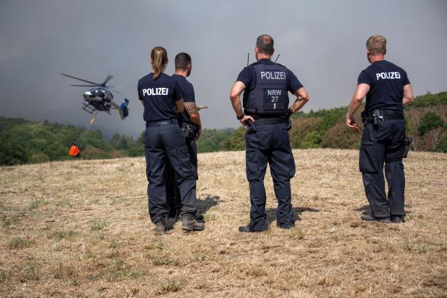 14 August 2026, North Rhine-Westphalia, Huertgenwald-Grosshau: Police officers stand on the scene at the wildfire in the Huertgenwald, while a helicopter flies overhead in the background carrying water to extinguish the fire. Photo: Harald Tittel/dpa