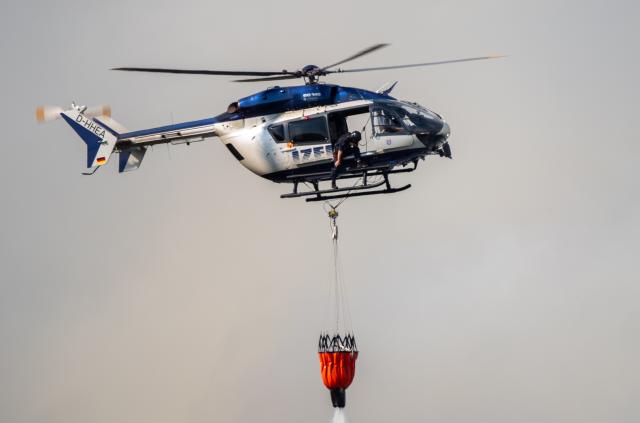14 August 2026, North Rhine-Westphalia, Huertgenwald-Grosshau: A police helicopter drops water from an external water tank onto a forest fire in the Hürtgenwald. Several hundred hectares of forest are ablaze. Photo: Harald Tittel/dpa