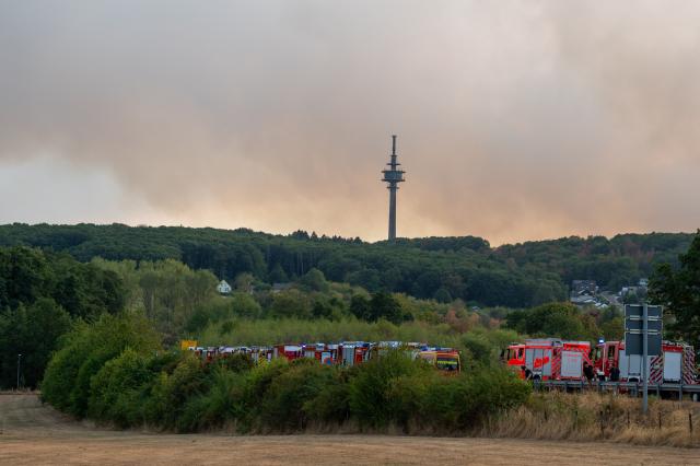 14 August 2026, North Rhine-Westphalia, Huertgenwald-Großhau: Firefighters wait on the B399 near Huertgenwald-Gey to respond to a wildfire in the Huertgenwald. In the background, the Grosshauer telecommunications tower is surrounded by clouds of smoke. Photo: Harald Tittel/dpa