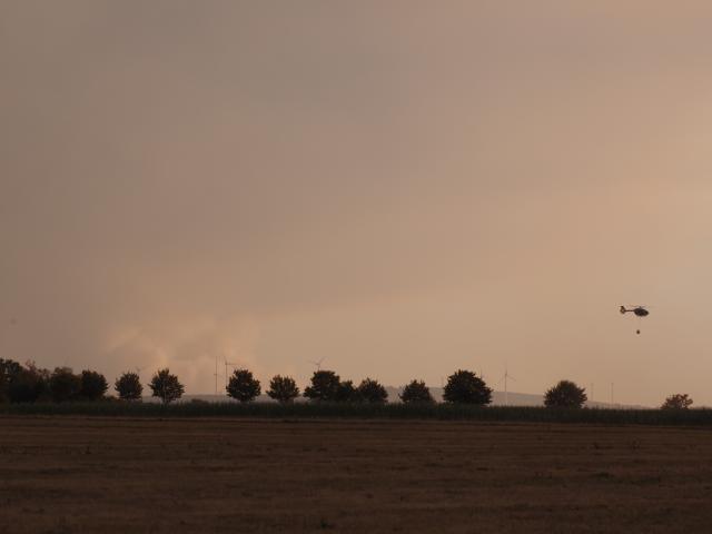 14 August 2026, North Rhine-Westphalia, Huertgenwald: A police helicopter equipped with a water tank flies over a forest fire in the municipality of Huertgenwald near Grosshau. Another large forest fire in the Hohen Venn can be seen in the distance. Photo: Georg Hilgemann/dpa