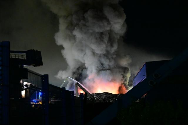 14 August 2026, North Rhine-Westphalia, Duisburg: Dark clouds of smoke rise into the night sky from a fire on a company property. In Duisburg, a pile of scrap metal measuring about 10,000 cubic meters is ablaze. Since Friday evening, emergency responders have been trying to extinguish the fire on a company property on the scrap island in the Port of Duisburg using water cannons and three excavators, said a fire department spokesperson. Photo: Alexander Forstreuter/dpa