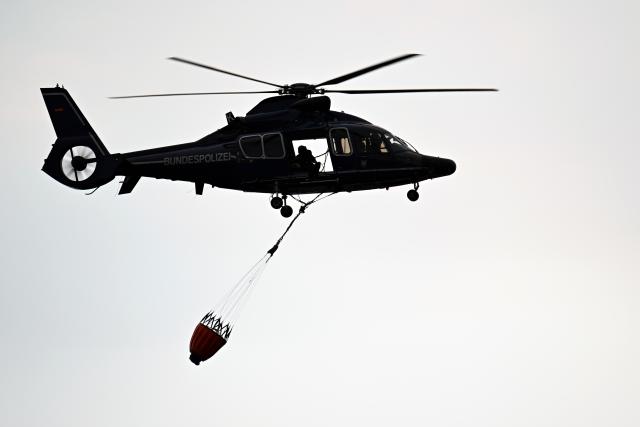 15 August 2026, North Rhine-Westphalia, Kleinhau: A police helicopter is flying over Kleinhau to collect water for the firefighting operation at the forest fire in the Huertgenwald. Several hundred hectares of forest are ablaze in the Huertgenwald forest fire. Photo: Federico Gambarini/dpa