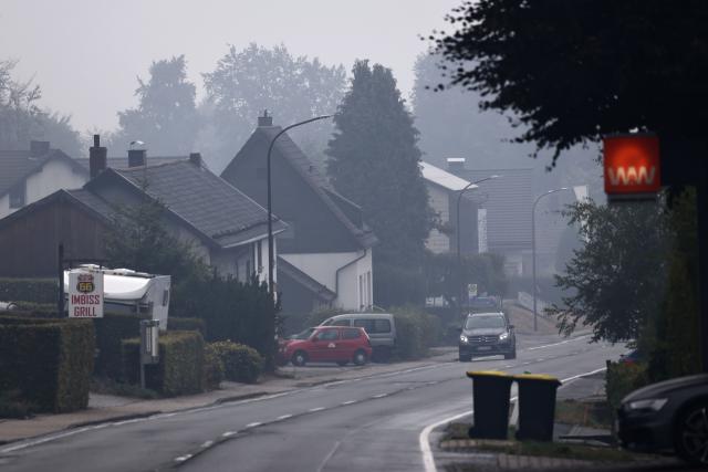 18 August 2026, North Rhine-Westphalia, Monschau: A car drives through the Mützenich district as plumes of smoke drift over the area affected by the fire. In the Hohes Venn nature reserve in Belgium, the battle against the wildfires continues. German emergency responders are also involved, operating out of the Eifel town of Monschau. Photo: Oliver Berg/dpa