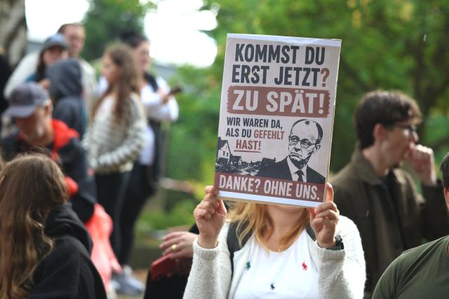 22 August 2026, North Rhine-Westphalia, Hürtgenwald: A woman holds a sign reading "Are you just getting here now? Too late!" during a visit by German Chancellor Friedrich Merz to Huertgenwald-Gey. Two days after the devastating wildfire in the Eifel region was completely extinguished, Chancellor Merz is visiting the area to thank the first responders. Photo: Christoph Reichwein/dpa