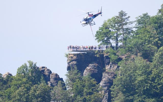 27 August 2026, Saxony, Hohnstein: A helicopter is on the scene clearing storm damage in Saxon Switzerland at Lake Amsel and near the Rathen Rock Theater; in the background, people are standing on the viewing platform at the Bastei. Photo: Robert Michael/dpa