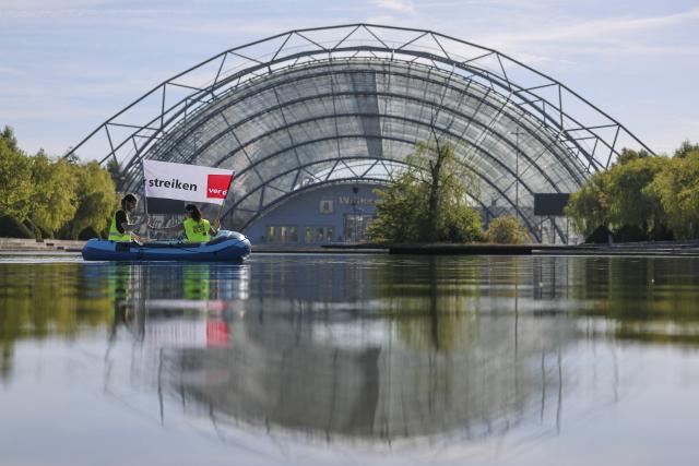 08 September 2026, Saxony, Leipzig: Employees of Leipziger Messe GmbH and its subsidiaries travel in a rubber boat carrying a banner that reads "We're on strike" across the lake in front of the Glass Hall at the exhibition grounds, as part of a warning strike. The union is demanding a pay raise that at least offsets inflation. Two rounds of collective bargaining have taken place so far. The third round of negotiations is scheduled for September 10. Photo: Jan Woitas/dpa