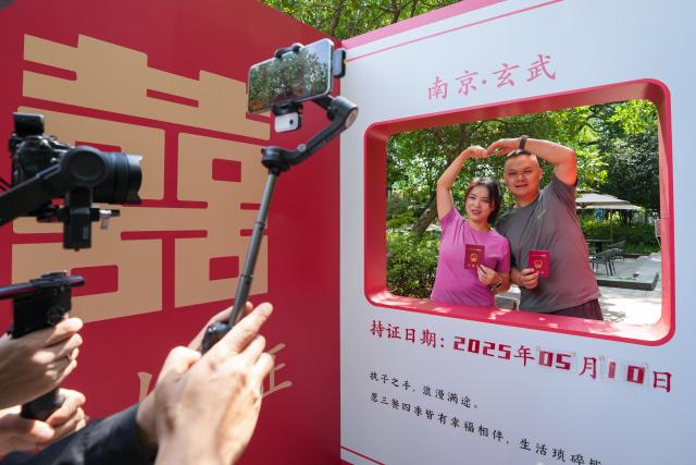 (250511) -- BEIJING, May 11, 2025 (Xinhua) -- A couple poses for photos with their marriage certificates at the Xuanwu Lake Park in Nanjing, east China's Jiangsu Province, May 10, 2025. (Xinhua/Li Bo)