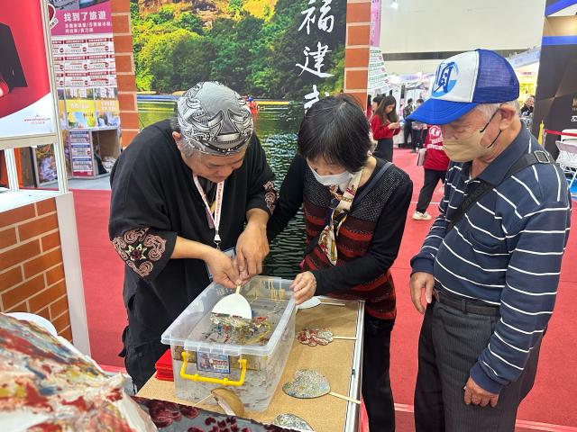 (251201) -- TAIPEI, Dec. 1, 2025 (Xinhua) -- This photo taken with a mobile phone shows lacquerware artist Huang Shou-kun (1st L) guiding a visitor to make a lacquer-colored miniature fan at the 2025 Kaohsiung Winter International Travel Fair in Kaohsiung, southeast China's Taiwan, Nov. 29, 2025. TO GO WITH "Fujian intangible cultural heritage draws visitors at Kaohsiung travel fair in Taiwan" (Xinhua/Huang Shuo)