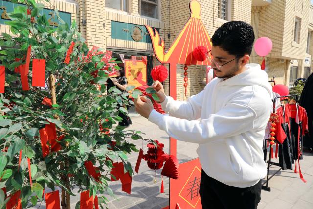 (260213) -- BAGHDAD, Feb. 13, 2026 (Xinhua) -- An Iraqi man hangs his New Year's wish on a tree on al-Mutanabbi Street in central Baghdad, Iraq, on Feb. 13, 2026. TO GO WITH "Feature: Red lanterns over Baghdad -- Chinese New Year celebration lights up Iraq's storied street of books" (Xinhua/Khalil Dawood)