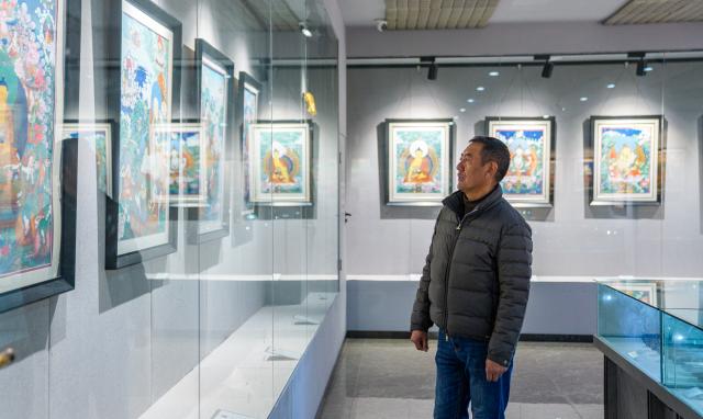 (260216) -- LHASA, Feb. 16, 2026 (Xinhua) -- Norbu Sidar looks at his own thangka works at the exhibition hall of the Xizang Academy of Thangka in Lhasa, southwest China's Xizang Autonomous Region, Feb. 10, 2026. On the afternoon of Feb. 10, sunlight streamed through the windows of the Xizang Academy of Thangka, illuminating an unfinished thangka painting. Norbu Sidar stood beside a young apprentice. He reached out, gently placing his finger on a specific spot on the canvas, and quietly guided the apprentice on the direction of the lines. 
   Thangka, renowned for its meticulous composition, vibrant colors, and profound cultural connotations, was listed among the first batch of China's national intangible cultural heritages in 2006. Recording Tibetan history, culture, medicine, astronomy, and other knowledge, it carries the spiritual beliefs and aesthetic wisdom of people in Xizang throughout the times.
   Norbu Sidar is a national-level inheritor of the Tibetan thangka. As a child, he learned thangka painting techniques from his grandfather. He recalled, "Time was most precious when I was little. I could only learn to paint thangka in my spare time after helping the family with farm and livestock work." From 2005 to 2015, he spent ten years as a restoration expert undertaking the mural restoration project at the Potala Palace. In 2016, he donated the 18 thangka works he had copied at the Potala Palace over the decade to the Palace. "These ten years have been the most meaningful part of my life," he recalled.
   In 1992, Norbu Sidar began taking on students. Ten years later, he led the founding of the Xizang Academy of Thangka. Today, as a master's supervisor at the School of Arts of Xizang University, he guides graduate students in studying the history of thangka and encourages young people to experiment with new forms of expression. To date, over 400 students are, like him, contributing to the development of Tibetan thangka art.
    After being elected as a member of the National Committee of the Chinese People's Political Consultative Conference, Norbu Sidar felt he had the responsibility to do more to make thangka, the artistic treasure in Xizang, recognized, supported and passed down through generations. (Xinhua/Tenzin Nyida)