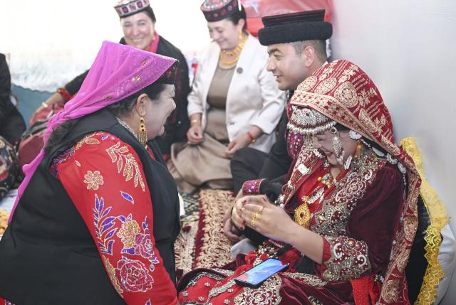 (260709) -- TAXKORGAN, July 9, 2026 (Xinhua) -- The groom's mother (L) chats with the bride at the wedding in Taxkorgan Tajik Autonomous County, northwest China's Xinjiang Uygur Autonomous Region, June 30, 2026. This summer, happiness filled the air around the home of herdsman Duri Jarman. His youngest son Nurkerim tied the knot with his beloved woman. Weddings of the Tajik ethnic group usually last three days. Inscribed on the national intangible cultural heritage list in 2008, the wedding customs of the Tajik ethnic group maintain unique traditions and are always described as festivals of music, dance and food to showcase the resplendent newlyweds and their propitious marriage.
  In Aimin Village, this wedding also serves as a vivid epitome of the eight-year transformative leap brought by the poverty alleviation relocation program.
  In the winter of 2018, Jarman and his family benefited from the relocation policy for poverty alleviation and settled in Aimin Village. They gradually adapted to the changes, enjoying a new life different from that of their ancestors. Jarman ran a small grocery store with Nurkerim, while his eldest and second sons both landed stable new jobs. Nurkerim also bought a pickup truck to run a transport business. It was this business that brought him together with his wife. For this family, the relocation is not only an opportunity for development, but also a path leading to happiness.
  Over the past eight years, Jarman has expanded his courtyard and planted saplings year after year. His small yard, where not a single tree used to grow, is now shaded by lush, thriving woods. As relocation sites of the poverty alleviation relocation program, Aimin Village and neighboring Fumin Village are now home to 321 households, with at least one person employed per household on average. (Xinhua/Hu Huhu)