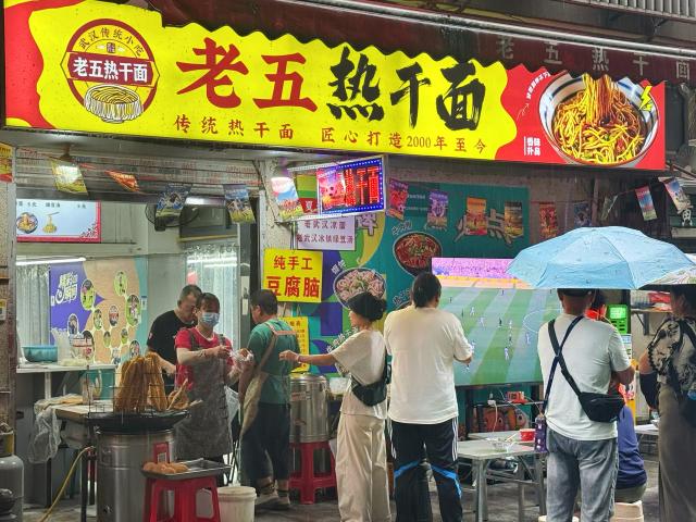 (260714) -- BEIJING, July 14, 2026 (Xinhua) -- Local residents and tourists enjoy a FIFA World Cup match in front of a breakfast shop in Wuhan, central China's Hubei Province, July 7, 2026. (Xinhua/Yu Xi)