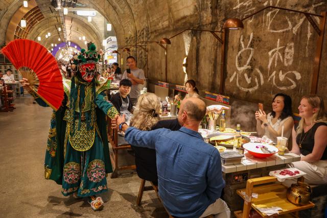 (260714) -- CHONGQING, July 14, 2026 (Xinhua) -- A foreign tourist interacts with a Sichuan Opera performer at a hotpot restaurant housed in a former underground air-raid shelter in Liangjiang New Area of southwest China's Chongqing Municipality, July 10, 2026. Since the beginning of this year, inbound and outbound passenger traffic at Chongqing Jiangbei International Airport has continued to grow, with a significant increase in foreign passenger traffic. In the first half of 2026, the airport handled more than 1.48 million inbound and outbound passenger trips, up 39.5 percent year on year. (Xinhua/Huang Wei)