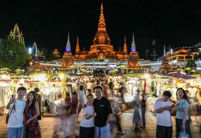 (260715) -- XISHUANGBANNA, July 15, 2026 (Xinhua) -- Tourists pose for photos at the Starlight Night Market in Jinghong, Xishuangbanna Dai Autonomous Prefecture, southwest China's Yunnan Province, July 14, 2026. The Starlight Night Market, located in Jinghong of Xishuangbanna Dai Autonomous Prefecture, has witnessed a surge in visitor arrivals during the summer peak tourism season. Featuring local delicacies, ethnic culture and diverse nighttime consumption options, this night market reflects the vitality of local cultural and tourism market. (Xinhua/Chen Xinbo)