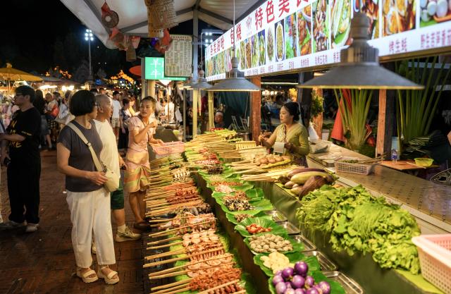 (260715) -- XISHUANGBANNA, July 15, 2026 (Xinhua) -- Tourists shop for local delicacies at the Starlight Night Market in Jinghong, Xishuangbanna Dai Autonomous Prefecture, southwest China's Yunnan Province, July 14, 2026. The Starlight Night Market, located in Jinghong of Xishuangbanna Dai Autonomous Prefecture, has witnessed a surge in visitor arrivals during the summer peak tourism season. Featuring local delicacies, ethnic culture and diverse nighttime consumption options, this night market reflects the vitality of local cultural and tourism market. (Xinhua/Chen Xinbo)