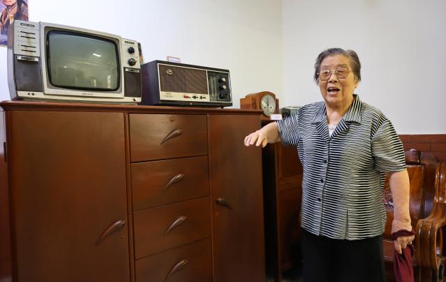 (260715) -- SHANGHAI, July 15, 2026 (Xinhua) -- Yu Meixiang recalls her life at her old residence in front of old household items displayed at a "time hall" in Pengyi residential compound, Jing'an District, east China's Shanghai, July 14, 2026. In Shanghai's Pengpu Xincun, one of the city's earliest newly built residential areas for in-service staff, senior residents like 92-year-old Yu Meixiang and 75-year-old Peng Juhua shared their memories of bygone days at a gathering with children of the neighborhood. Their stories reflect not only personal history but also a remarkable urban renewal that has transformed their lives while keeping their community intact.
   Built mostly in the 1950s and 1960s, the residential buildings in Pengpu Xincun had long fallen into disrepair, as could be seen in the leaky roofs and crumbling walls. The daily hardships had been worsened by kitchens and bathrooms shared among neighbors, especially when families there were growing in sizes. 
   In response, the local government launched a renewal project in 2005. Residents temporarily relocated while their old homes were reconstructed on the original plots. After two decades of work in multiple phases, Pengyi residential compound of Pengpu Xincun officially opened for its earlier residents to move back in June 2025.
   The refurbished apartments there are now more spacious, each with a private kitchen and bathroom. Elevators, barrier-free access, and a pedestrian-friendly internal traffic system are now in place to make life much easier for the elderly. A neighborhood cultural center with a library and gym is set to open soon. (Xinhua/Fang Zhe)