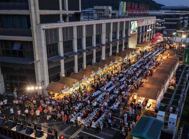 (260725) -- JIANDE, July 25, 2026 (Xinhua) -- An aerial drone photo taken on July 24, 2026 shows people enjoying foods and drinks and watching a performance at a beer festival in Jiande City of east China's Zhejiang Province. A summer beer festival kicked off at a farmers market here on Friday. Featuring live performances, gourmet foods and drinks, leisure activities and bazaars, the event attracted thousands of locals and tourists. (Xinhua/Xu Yu)