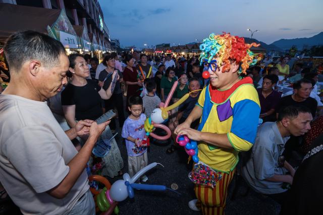 (260725) -- JIANDE, July 25, 2026 (Xinhua) -- An artist interacts with visitors at a beer festival in Jiande City of east China's Zhejiang Province on July 24, 2026. A summer beer festival kicked off at a farmers market here on Friday. Featuring live performances, gourmet foods and drinks, leisure activities and bazaars, the event attracted thousands of locals and tourists. (Xinhua/Xu Yu)