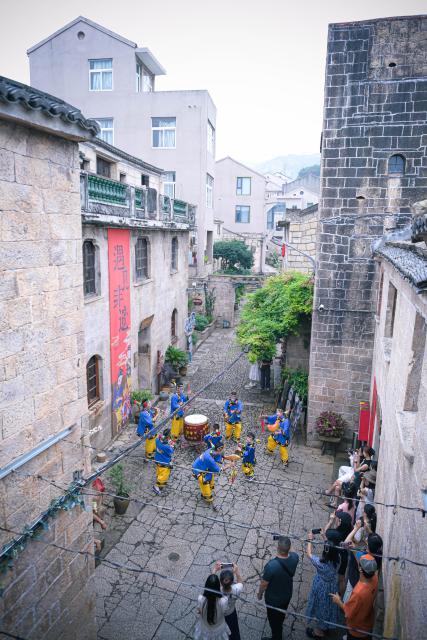 (260726) -- WENLING, July 26, 2026 (Xinhua) -- Tourists watch a performance of Dazou drum, a national intangible cultural heritage,  in Liruo Village of Shitang Town, Wenling City, east China's Zhejiang Province, June 28, 2026. Liruo is a village located in the southwestern part of Shitang Town, Wenling City. For generations, local residents have relied on the sea for their livelihood. The village is known for its distinctive stone constructions, which were built to withstand typhoons and other severe weather.
   Today, the ancient fishing village is boosting its cultural and tourism industries. Many traditional stone houses have been transformed into cafes and homestays, while fishing nets are crafted into handbags, serving as souvenirs for tourists together with other cultural and creative products. (Xinhua/Jiang Han)