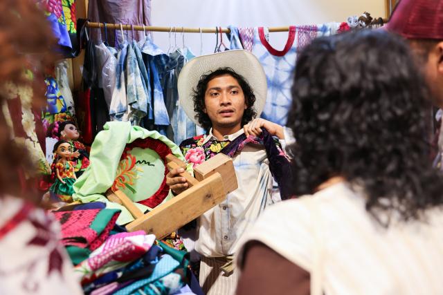 (260731) -- OAXACA, July 31, 2026 (Xinhua) -- An artisan shows a hand-embroidered garment to customers at the 2026 Guelaguetza Handicrafts Fair in Oaxaca City, Oaxaca State, Mexico, July 26, 2026. The southern Mexican state of Oaxaca is known for its handwoven textiles and embroidered garments. Local artisans make clothing, rugs and other items using weaving, dyeing and embroidery techniques. Sold at markets and handicraft fairs, these colorful creations remain part of everyday life in Oaxaca. (Xinhua/Liu Wenhui)
