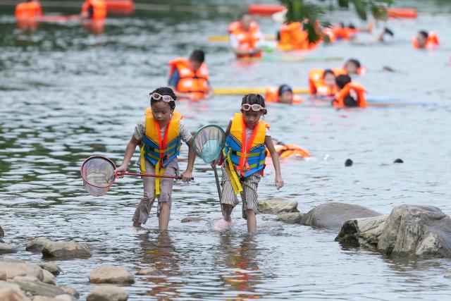 (260806) -- ANJI, Aug. 6, 2026 (Xinhua) -- Children catch fish in a stream in Anji County, east China's Zhejiang Province, Aug. 6, 2026. During the summer vacation, Anji County leverages its cool weather and beautiful scenery, attracting numerous tourists to escape the summer heat. (Xinhua/Huang Zongzhi)