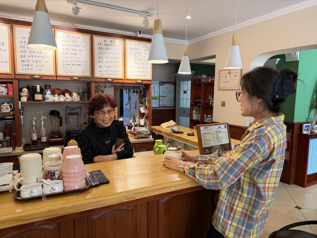 (260807) -- QINGDAO, Aug. 7, 2026 (Xinhua) -- Zhao Qiying (L) talks with a customer at Rona Coffee in Qingdao, east China's Shandong Province, June 15, 2026. TO GO WITH "Feature: Cafe's success serves to bring wider world to 86-year-old proprietor" (Xinhua/Zhang Liyuan)
