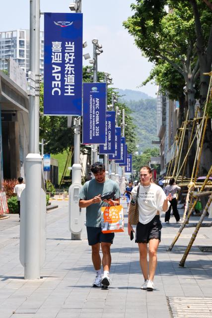 (260810) -- SHENZHEN, Aug. 10, 2026 (Xinhua) -- Foreign tourists walk past themed banners on a street in Huaqiangbei of Shenzhen, south China's Guangdong Province, Aug. 10, 2026. With themed bulletin boards, outdoor posts, decorations and installations, Shenzhen marked on Monday the 100-day countdown to the 33rd Asia-Pacific Economic Cooperation (APEC) Economic Leaders' Meeting. (Xinhua/Liang Xu)