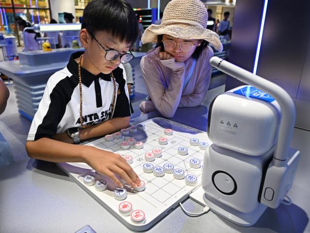 (260811) -- QINGDAO, Aug. 11, 2026 (Xinhua) -- A boy plays Chinese chess with a robot at a robot 6S experience store in Qingdao, east China's Shandong Province on Aug. 11, 2026. Recently, the first robot 6S experience store in Qingdao opened, attracting a large number of consumers to visit and experience. This store integrates six major functions including sale, spare parts, service, survey, lease and personalized customization, helping further introducing robots into people's lives. (Xinhua/Li Ziheng)