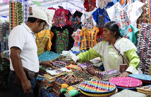 (260819) -- MEXICO CITY, Aug. 19, 2026 (Xinhua) -- A vendor introduces embroidered garments and woven products to a visitor during the 12th Indigenous Cultures Festival at Constitution Square in Mexico City, capital of Mexico, Aug. 18, 2026. Inside a large exhibition tent, visitors browse goods and taste local specialty drinks, while artisans demonstrate traditional skills such as embroidery. The festival will run until Aug. 23. (Xinhua/Liu Wenhui)