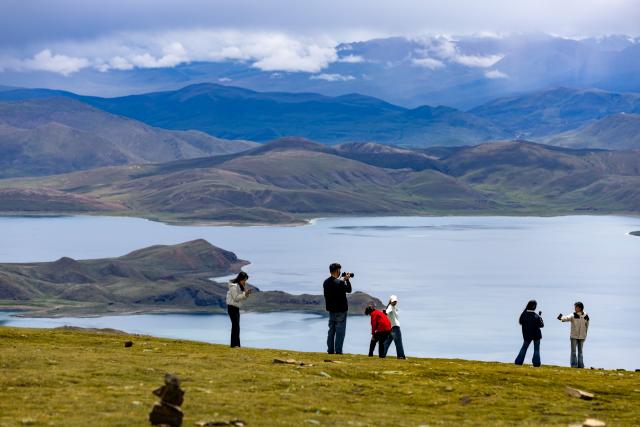 (260821) -- LHASA, Aug. 21, 2026 (Xinhua) -- Tourists pose for photos with Lake Yamzhog Yumco, Shannan City, southwest China's Xizang Autonomous Region, Aug. 20, 2026. (Xinhua/Jiang Fan)