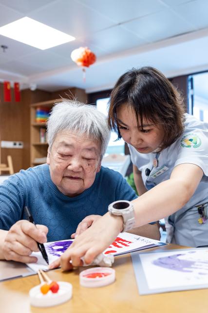 (260823) -- HUZHOU, Aug. 23, 2026 (Xinhua) -- Shen Wanting (R) helps a senior resident make handicrafts in Huzhou City, east China's Zhejiang Province, Aug. 17, 2026.
  Senior residents at the Shushan Healthcare Center, an elderly care center in Huzhou, Zhejiang Province, sometimes misunderstand one another during daily conversations because of their advanced age. These moments inspired Shen Wanting, a post-95 nurse, and five colleagues of similar age to create micro dramas featuring these elderly residents and their daily lives at the center. 
  Five senior residents, whose average age is over 80, are regular cast members, and the oldest is over 90. 
  After gaining attention online, the production team has continued to release micro drama episodes. Since June 2025, they have produced over 160 episodes. (Photo by Yi Fan/Xinhua)