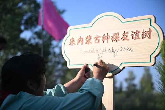 (260824) -- ORDOS, Aug. 24, 2026 (Xinhua) -- Yin Yuzhen signs her name after planting a tree in the Maowusu Sandy Land in north China's Inner Mongolia Autonomous Region, Aug. 24, 2026. Retired U.S. teacher Ronald Sakolsky revisited the Maowusu Sandy Land on Monday and planted sapling with his friend Yin Yuzhen, a national model worker who has been planting trees here for four decades, as well as Yin's husband Bai Wanxiang.
   Their story began in 1999, when Sakolsky taught English at a school in central China's Henan Province under a teachers' exchange program between the two countries.
   After watching a television news report featuring Yin's tireless work, he was deeply moved and raised 5,000 U.S. dollars to help Yin and her husband in their afforestation efforts against desertification.
   The saplings bought and planted with that donation have since grown into a forest of more than 50,000 towering trees. (Xinhua/Lian Zhen)