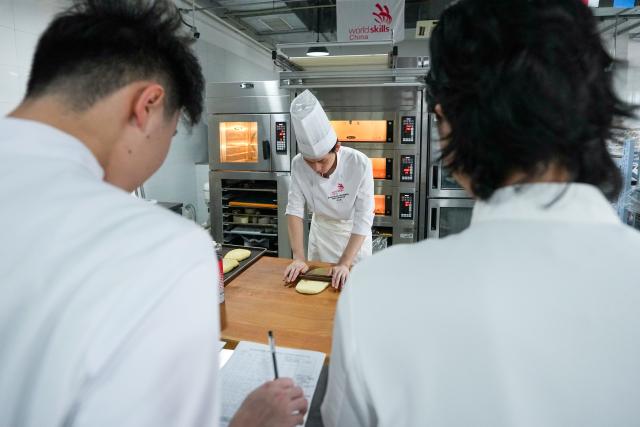 (260919) -- SUZHOU, Sept. 19, 2026 (Xinhua) -- Hou Jinchen (C) makes bread based on his training plan at Suzhou Wuzhong Technician College in Suzhou, east China's Jiangsu Province, Aug. 27, 2026. Born in 2005, Hou Jinchen studies culinary arts at Suzhou Wuzhong Technician College. He began learning to bake at the age of 12, and his passion for it has grown ever since.
   From shaping dough to baking and decorating bread, Hou follows a rigorous training routine. He practices the same techniques over and over to build muscle memory, carefully recording changes in temperature and ingredient quantities during each session and constantly refining the details.
   Outside training, he also visits local bakeries to learn from working bakers. He believes they have a better understanding of consumer preferences, which gives him insights that training focused solely on competitions may not always provide.
   His efforts have earned him both gold and bronze medals in national vocational skills competitions, and he was later selected for the national team. The 21-year-old is now preparing for the upcoming WorldSkills Competition in Shanghai.
   Among all the competition tasks, Hou devotes particular attention to artistic bread design. From sketching ideas to making molds and baking the final pieces, the process requires repeated adjustments. He sometimes uses 3D-printed molds and plans to incorporate more Chinese elements into his competition pieces in Shanghai.
   For Hou, baking is not just a technical skill, but also a pursuit of craftsmanship. He hopes to showcase the skills and dedication of young Chinese bakers on the international stage.
   "I can't imagine leaving the baking industry," Hou said. "In the future, I hope to become a coach and pass on what I have learned to younger bakers." (Xinhua/Li Bo)