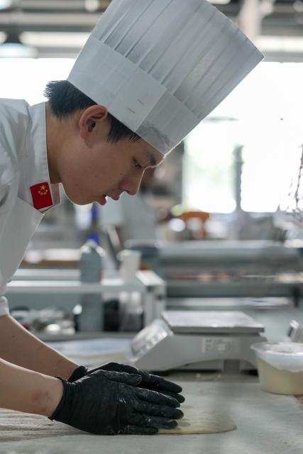 (260919) -- SUZHOU, Sept. 19, 2026 (Xinhua) -- Hou Jinchen makes bread based on his training plan at Suzhou Wuzhong Technician College in Suzhou, east China's Jiangsu Province, Aug. 27, 2026. Born in 2005, Hou Jinchen studies culinary arts at Suzhou Wuzhong Technician College. He began learning to bake at the age of 12, and his passion for it has grown ever since.
   From shaping dough to baking and decorating bread, Hou follows a rigorous training routine. He practices the same techniques over and over to build muscle memory, carefully recording changes in temperature and ingredient quantities during each session and constantly refining the details.
   Outside training, he also visits local bakeries to learn from working bakers. He believes they have a better understanding of consumer preferences, which gives him insights that training focused solely on competitions may not always provide.
   His efforts have earned him both gold and bronze medals in national vocational skills competitions, and he was later selected for the national team. The 21-year-old is now preparing for the upcoming WorldSkills Competition in Shanghai.
   Among all the competition tasks, Hou devotes particular attention to artistic bread design. From sketching ideas to making molds and baking the final pieces, the process requires repeated adjustments. He sometimes uses 3D-printed molds and plans to incorporate more Chinese elements into his competition pieces in Shanghai.
   For Hou, baking is not just a technical skill, but also a pursuit of craftsmanship. He hopes to showcase the skills and dedication of young Chinese bakers on the international stage.
   "I can't imagine leaving the baking industry," Hou said. "In the future, I hope to become a coach and pass on what I have learned to younger bakers." (Xinhua/Li Bo)